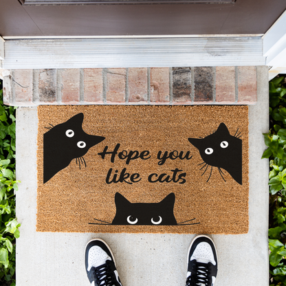 Hope You Like Cats coir doormat with three peeking black cats in front of a brick step, top-down view with sneakers