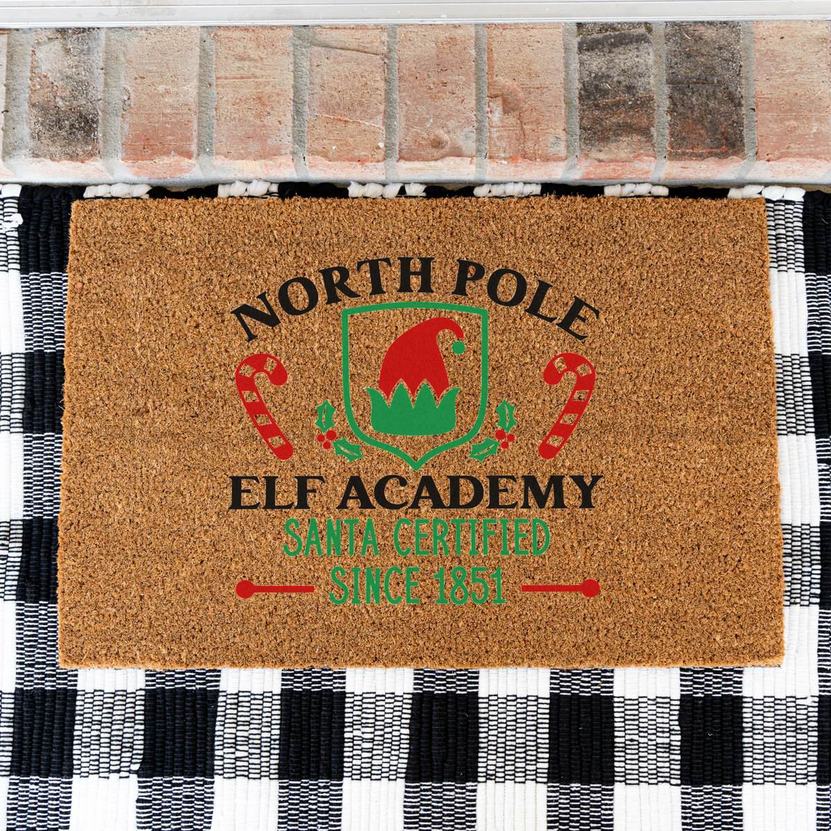 North Pole Elf Academy coir doormat on a black and white buffalo check rug, close view