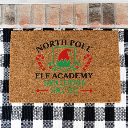 North Pole Elf Academy coir doormat on a black and white buffalo check rug, close view