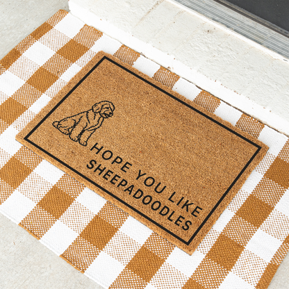 Hope You Like Sheepadoodles coir doormat with a drawing of a sitting Sheepadoodle at an angle on a tan and white check rug