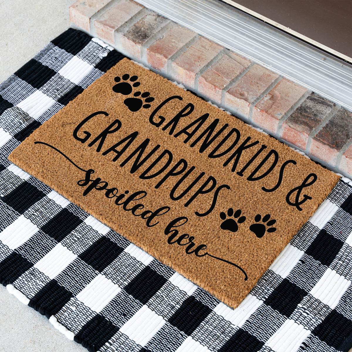 Grandkids & Grandpups Spoiled Here coir doormat with paw prints at an angle on a black and white buffalo check rug