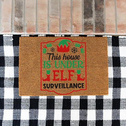 This House Is Under Elf Surveillance framed coir doormat on a black and white buffalo check rug at a brick step
