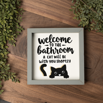 Welcome to the bathroom, a cat will be with you shortly framed sign with a grey frame on a dark wood table with greenery