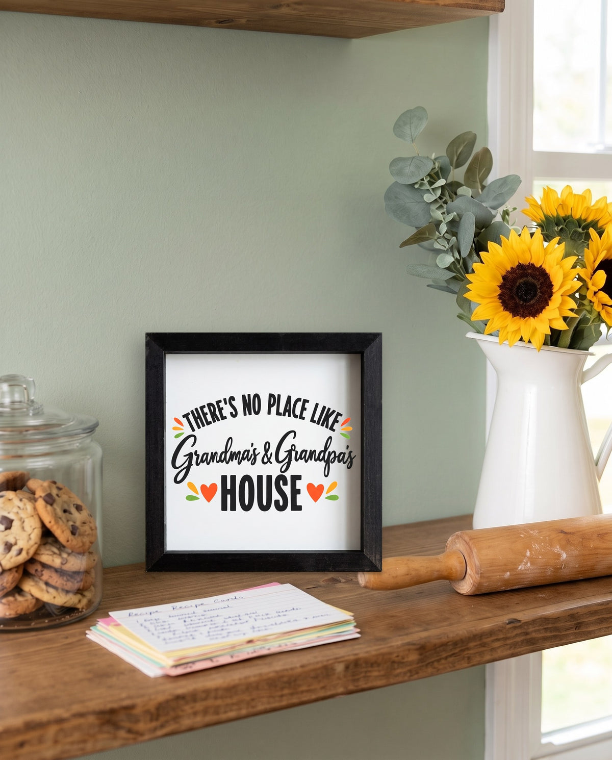 There's No Place Like Grandma's & Grandpa's House square framed sign with a black frame, shown in 12x12, on a kitchen shelf beside cookies and sunflowers