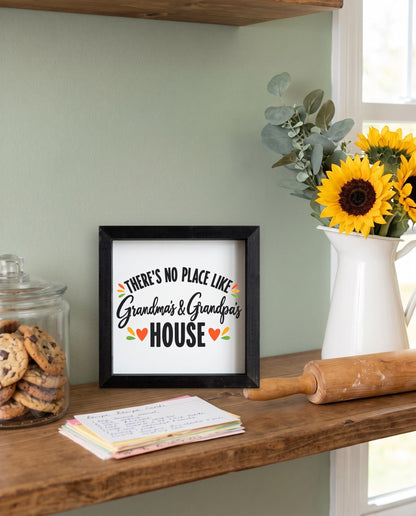 There's No Place Like Grandma's & Grandpa's House square framed sign with a black frame, shown in 12x12, on a kitchen shelf beside cookies and sunflowers